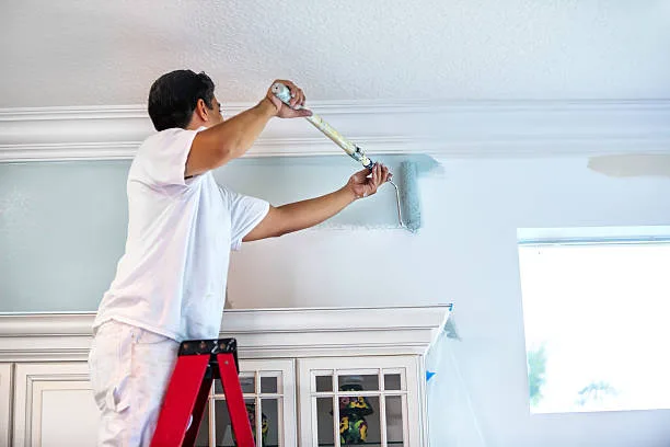 A housepainter stands on a ladder to paint the walls inside a residential home.  rr