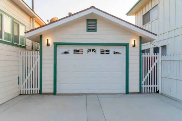 Detached garage in between two white buildings at Oceanside, California. Garage exterior with white sectional door with green frames and two wall lamps.