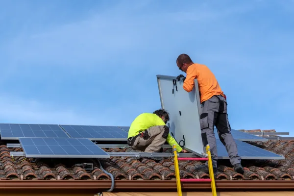 Roof with two people installing solar panels and a blue sky with white clouds in the background.